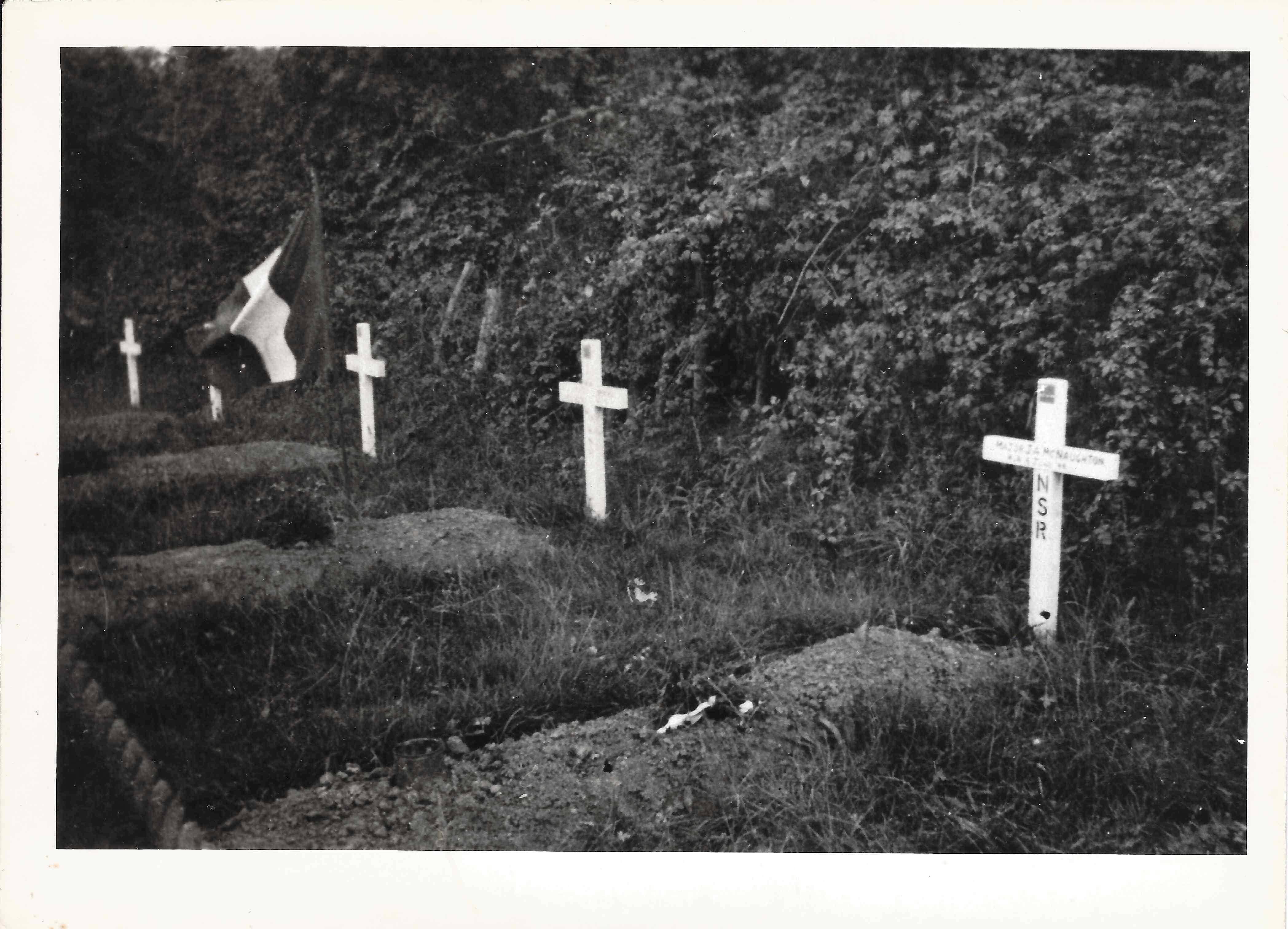 Photographie en noir et blanc – Une croix blanche est dressée à la tête d’une tombe venant d’être creusée, parmi d’autres croix blanches et près d’un drapeau français. Les tombes se trouvent sous plusieurs arbres. Un bouquet a été posé sur le dessus de ch